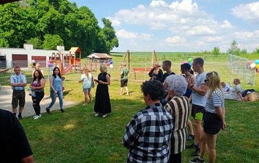 Na zdjęciu widoczna jest koordynatorka projektu Anna Olejniczak oraz mieszkańcy słuchający przem&oacute;wienia W&oacute;jta Gminy Rąbino Anety Krawiec. Za zgromadzonymi znajduje się nowo powstałe ogrodzenie placu zabaw na rzecz Rewitalizacji Dąbrowy Białogardzkiej.